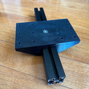 May include: A black T-slot aluminum extrusion rail attached to a rectangular black mounting plate. The plate features a grid of pre-drilled holes and a central silver metal fastener, forming a structural component for mechanical assembly.