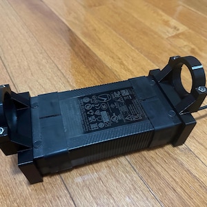 Könnte beinhalten: Schwarzer Netzadapter mit rechteckiger Form und einem runden Stecker an jedem Ende. Der Adapter hat ein Etikett mit Text und Symbolen, darunter "E11 UK CE".