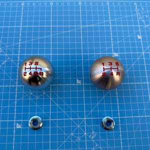 Pode incluir: Dois botões de alavanca de câmbio prateados com padrões de mudança de marcha de 5 velocidades vermelhos. Os botões estão em um fundo de grade azul.