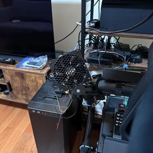 May include: A black computer case with a mounted fan and a black metal frame. The fan has a honeycomb grill. Wires in yellow, red, and white are visible. The setup is near a black monitor and a wooden cabinet.
