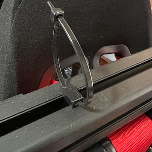 Puede incluir: Una brida negra que asegura un componente, probablemente parte de un asiento de vehículo. La brida está en bucle y sujeta, con un respaldo negro y un cinturón de seguridad rojo visibles en el fondo.