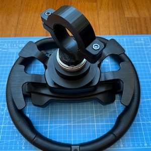 May include: A black racing steering wheel with a black quick release hub and a black clamp. The steering wheel is on a blue mat.