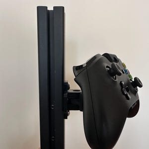 May include: A black video game controller is mounted to a black vertical stand. The controller is attached to the stand with a black plastic bracket.