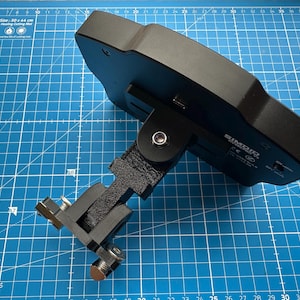 May include: A black electronic device with the text "SIMAIR" and a power switch, mounted on a black bracket with a fabric strap and metal hardware. The device is on a blue cutting mat with grid lines and the text "Elan Arts & Crafts".