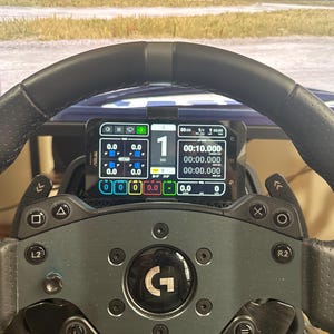 Può includere: Un volante da corsa nero con un centro grigio e un supporto per telefono nero e bianco fissato in alto. Il supporto del telefono mostra un'app di corsa con un timer, i tempi sul giro e altri dati. Il volante ha pulsanti e paddle per controllare il gioco.