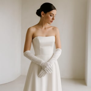 Könnte beinhalten: Ein trägerloses weißes Brautkleid aus Satin mit weitem Rock. Die Braut trägt lange, weiße Satinhandschuhe, die mit kleinen, gleichmäßig verteilten Perlen verziert sind. Die Handschuhe reichen über die Ellbogen hinaus. Das Haar der Braut ist zu einem tiefen Dutt frisiert.