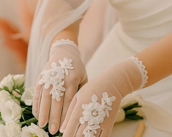 Könnte beinhalten: Elfenbeinfarbene, transparente Handschuhe mit floralen Spitzenapplikationen. Die Handschuhe sind mit aufwendigen weißen Spitzenblumen und Perlen verziert. Das Bild zeigt auch einen Strauß weißer Blumen und den Text "Lydia SUMMER BLOOM".