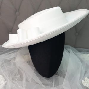 Peut inclure: Chapeau blanc avec un large bord et une couronne cylindrique. Le chapeau est orné d'un ruban blanc et de trois nœuds décoratifs blancs. Il est présenté sur une tête de mannequin noire, avec un tissu blanc transparent en arrière-plan.