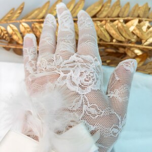 Op de afbeelding: Close-up van een witte kanten handschoen met een bloemmotief. De handschoen is versierd met veren en een satijnen lint. De achtergrond toont een gouden bladerarrangement, wat een delicate en elegante esthetiek creëert.