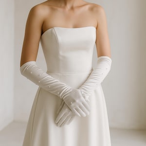 Op de afbeelding: Een witte strapless trouwjurk met lange, doorschijnende handschoenen versierd met kleine parels. De handschoenen reiken tot voorbij de ellebogen, wat elegantie toevoegt. De jurk heeft een eenvoudig, vloeiend ontwerp, perfect voor een bruiloft.