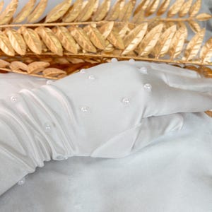 Op de afbeelding: Witte, doorschijnende handschoenen versierd met kleine, gelijkmatig verdeelde parel kralen. De handschoenen zijn gemaakt van een delicate, vloeiende stof en worden getoond tegen een achtergrond van goudkleurige bladeren.