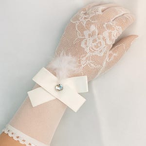 Op de afbeelding: Een crèmekleurige kanten handschoen met een bloemmotief. Een witte satijnen strik met een kristal en een kleine veer sieren de pols. De handschoen heeft een delicaat, vrouwelijk ontwerp, ideaal voor evenementen.