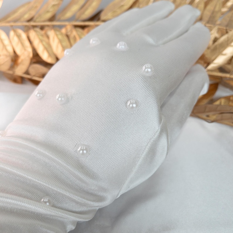 Op de afbeelding: Een witte satijnen handschoen met kleine, gelijkmatig verdeelde parelversieringen. De handschoen is gemaakt van een gladde, licht glanzende stof. De parels zijn rond en geven het accessoire een vleugje elegantie.