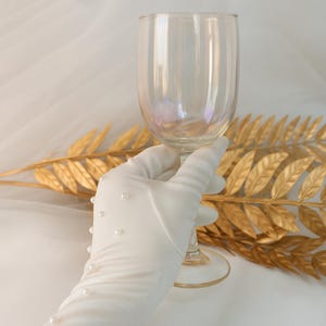 Op de afbeelding: Een helder iriserend wijnglas wordt vastgehouden door een witte handschoen versierd met kleine parels. Het glas reflecteert licht, waardoor een regenboogeffect ontstaat. Een gouden decoratieve bladertak staat op de achtergrond.