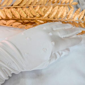 Op de afbeelding: Een witte satijnen handschoen versierd met kleine, gelijkmatig verdeelde parels. De handschoen staat tegen een achtergrond van goudkleurig blad, wat een contrast van texturen en kleuren cre&euml;ert. De stof van de handschoen heeft een subtiele glans.