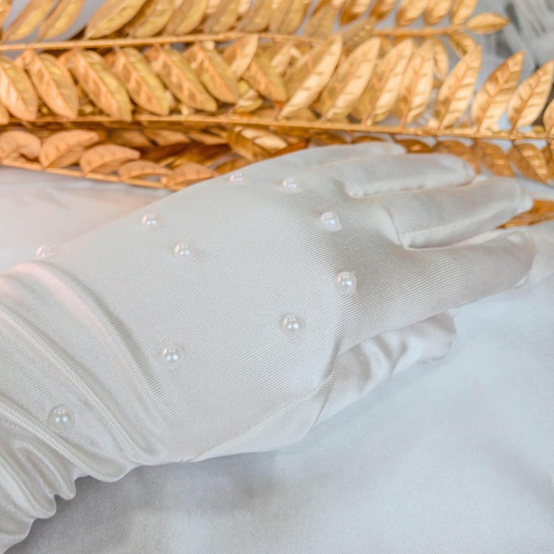 Op de afbeelding: Een witte satijnen handschoen versierd met kleine, gelijkmatig verdeelde parels. De handschoen staat tegen een achtergrond van goudkleurig blad, wat een contrast van texturen en kleuren cre&euml;ert. De stof van de handschoen heeft een subtiele glans.