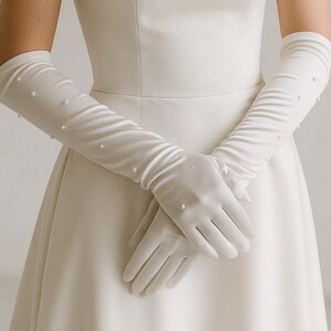 Op de afbeelding: Elegante witte satijnen handschoenen met parelaccenten. De lange handschoenen reiken tot voorbij de ellebogen en worden gecombineerd met een witte jurk. De handschoenen hebben een glad, vloeiend ontwerp.