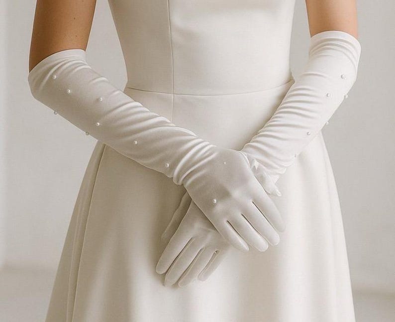 Op de afbeelding: Elegante witte satijnen handschoenen met parelaccenten. De lange handschoenen reiken tot voorbij de ellebogen en worden gecombineerd met een witte jurk. De handschoenen hebben een glad, vloeiend ontwerp.