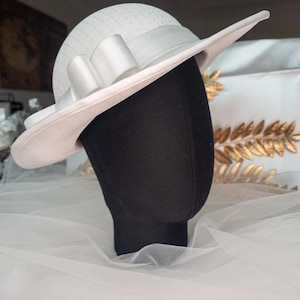 May include: A white hat with a light pink brim and a white ribbon bow. The hat has a textured crown and a wide brim. The hat is displayed on a black mannequin head.