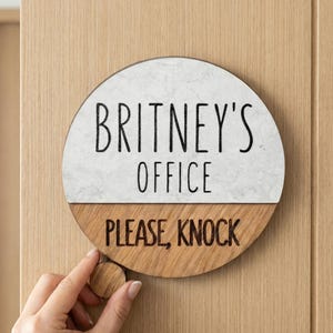 Puede incluir: Un letrero de oficina redondo con el texto "BRITNEY'S OFFICE PLEASE, KNOCK". El letrero es de dos tonos, con una parte superior blanca con aspecto de mármol y una parte inferior de madera. Se adjunta un pequeño pomo de madera.