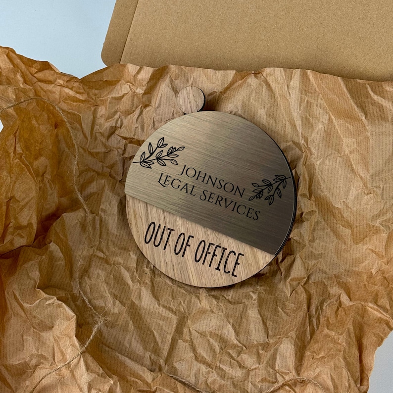 Pu&ograve; includere: Un cartello in legno con un design diviso. La met&agrave; superiore &egrave; in legno marrone chiaro con il testo "JOHNSON LEGAL SERVICES" e un motivo a foglie. La met&agrave; inferiore &egrave; in legno marrone scuro con il testo "OUT OF OFFICE".