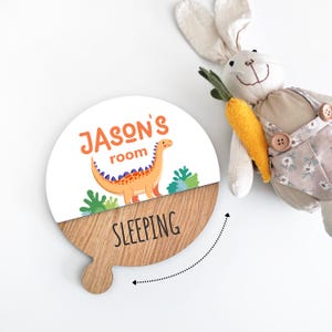 May include: A round wooden sign with the text "JASON'S room" and a cartoon dinosaur illustration. The bottom half of the sign is wood, with the word "SLEEPING" printed on it. A stuffed bunny with a carrot is in the background.