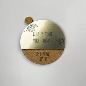 Pu&ograve; includere: Un cartello rotondo in legno con una met&agrave; superiore in metallo dorato. La met&agrave; superiore ha il testo "What's your vibe today?" e un piccolo disegno floreale. La met&agrave; inferiore &egrave; in legno e ha il testo "I'm feeling chatty".