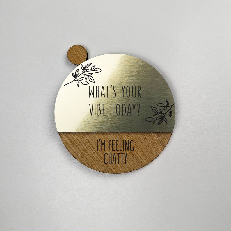 Pu&ograve; includere: Un cartello rotondo in legno con una met&agrave; superiore in metallo dorato. La met&agrave; superiore ha il testo "What's your vibe today?" e un piccolo disegno floreale. La met&agrave; inferiore &egrave; in legno e ha il testo "I'm feeling chatty".