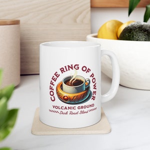 May include: White ceramic mug with a red and gold design. The mug features a coffee cup with steam rising from it, inside a gold ring with a red jewel. The text "Coffee Ring of Power" is written in a circle around the cup. The text "Volcanic Ground Dark Roast Blend" is written below the cup.