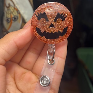 May include: A round orange glitter badge reel featuring a black jack-o'-lantern face with prominent eyelashes. The circular resin topper is attached to a clear plastic strap with a metal snap button and a small metal swivel clip for attachment.