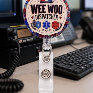 May include: A round badge reel featuring a red, white, and blue glitter border. The center graphic displays the text "WEE WOO DISPATCHER" with icons of a light bar, a radio, a star of life, and a fire department emblem in red, white, and blue.