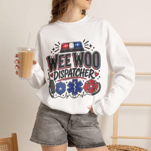 May include: A white long-sleeve sweatshirt featuring a graphic print with the text WEE WOO DISPATCHER in black and red. The design includes a police light bar, a radio, a police badge, a medical star of life, and a fire department emblem.