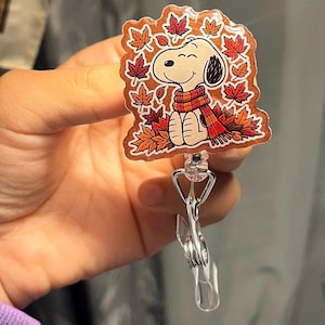 May include: A badge reel featuring a cartoon dog with black ears wearing a red and orange plaid scarf, surrounded by falling autumn maple leaves in shades of red, orange, and yellow. The design is set against a glittery copper-toned background.