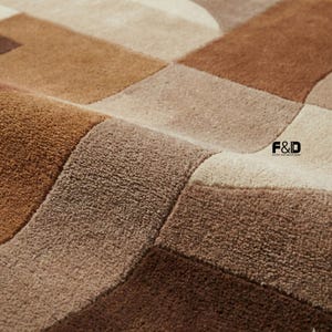 Könnte beinhalten: Nahaufnahme eines geometrischen Teppichs in Braun-, Beige- und Cremetönen. Der Teppich hat ein weiches, strukturiertes Aussehen mit einem sich wiederholenden Muster aus gebogenen und rechteckigen Formen. Der Markenname "F&D" ist sichtbar.