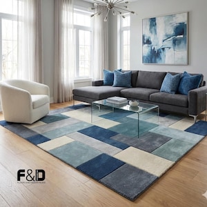Könnte beinhalten: Ein modernes Wohnzimmer mit einem grauen Ecksofa, einem weißen Sessel und einem Glastisch. Ein großer Teppich mit einem geometrischen Muster in Blau-, Grau- und Cremetönen bedeckt den Boden. Ein Gemälde mit blauer abstrakter Kunst hängt an der Wand.
