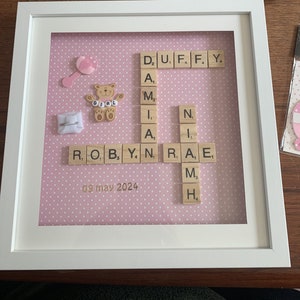 May include: A pink and white framed picture with a teddy bear and a baby rattle. The frame contains a scrabble tile word art with the names Robyn and Duffy and the date 09 may 2024.