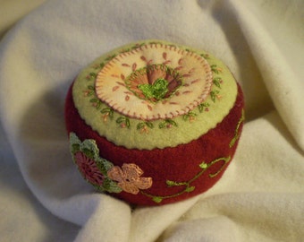 May include: A hand-stitched pincushion shaped like an apple with a green and white top and a red bottom. The top features a circular design with a flower in the center.