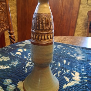 May include: A tall ceramic vase with a flared base and a narrow neck. The lower portion is a muted blue-gray, while the upper section features a textured, dark brown band with repeating vertical and horizontal etched patterns in the clay surface.