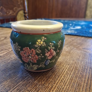 May include: A small, rounded ceramic planter with a white rim and a deep green body. The exterior features a hand-painted floral design with pink, yellow, and blue flowers and green leaves. The vessel has a smooth, glossy finish and a wide opening.