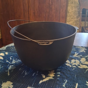 May include: A large, round, black cast iron pot with a smooth, deep basin and a curved metal bail handle attached to two small side lugs. The pot features a wide, open top and a rounded bottom, presenting a heavy, solid construction.