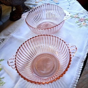 May include: Two matching pink glass bowls featuring a vertical ribbed pattern along the sides and a sunburst design on the interior base. Each bowl has two small integrated handles on opposite sides and a scalloped edge along the top rim.