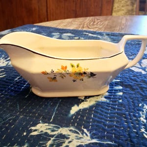 May include: A white ceramic gravy boat featuring a gold-trimmed rim and a floral pattern with yellow and orange blossoms and green leaves. The vessel has a curved pouring spout, a sturdy handle, and a faceted body design with a pedestal base.