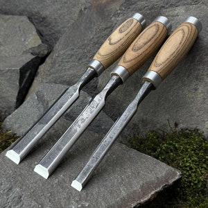 Puede incluir: Tres cinceles de mango de madera con hojas de metal, dispuestos en fila sobre una roca gris con musgo verde. Los cinceles son de diferentes tamaños, siendo el más grande el de abajo y el más pequeño el de arriba.