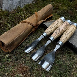 May include: Set of three wood carving tools with wooden handles and a leather roll-up case. The tools have a silver finish and are arranged on a green mossy surface.