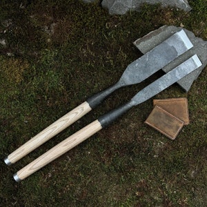 Puede incluir: Dos cinceles de metal con mango de madera y acabado gris, sobre un lecho de musgo verde. También se pueden ver dos cuadrados de cuero marrón en la imagen.