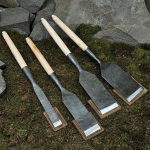 Puede incluir: Cuatro formones para carpintería con mangos de madera y hojas de metal. Los formones están dispuestos sobre un lecho de musgo verde y se colocan contra un fondo de rocas grises. Cada formón descansa sobre una pequeña almohadilla de cuero marrón.