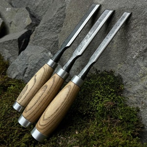 Puede incluir: Tres cinceles de mango de madera con hojas de metal, dispuestos sobre un lecho de musgo verde y rocas.