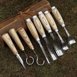May include: A set of eight wood carving tools with wooden handles and metal blades, rolled up in a brown leather case.