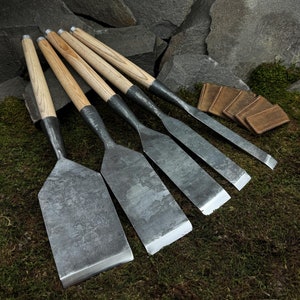 Puede incluir: Conjunto de seis cinceles de metal con mango de madera y acabado gris. Los cinceles están dispuestos en fila sobre un lecho de musgo verde.