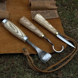 May include: Three wood-handled carving tools with metal blades are displayed on a brown leather roll-up pouch. The tools include a hook knife, a spoon knife, and a gouge.